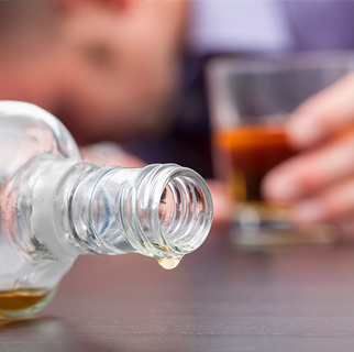 close up image of a spilled bottle of whisky, a glass with a hand on it and a mans face, face down on a table.